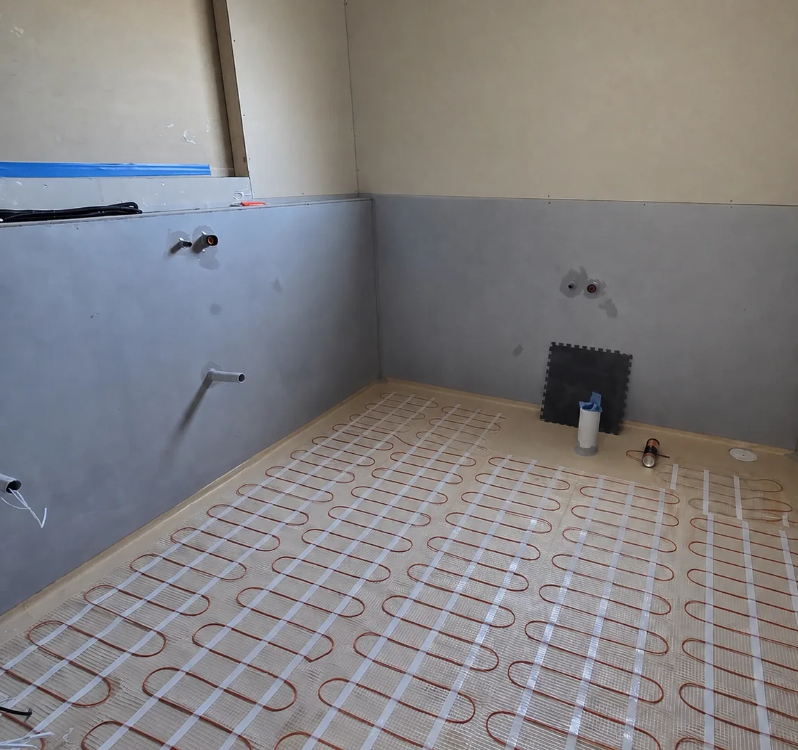 A heating mat laid across a bathroom floor, with the walls rendered and the pipe stubs left ready for the fitout.