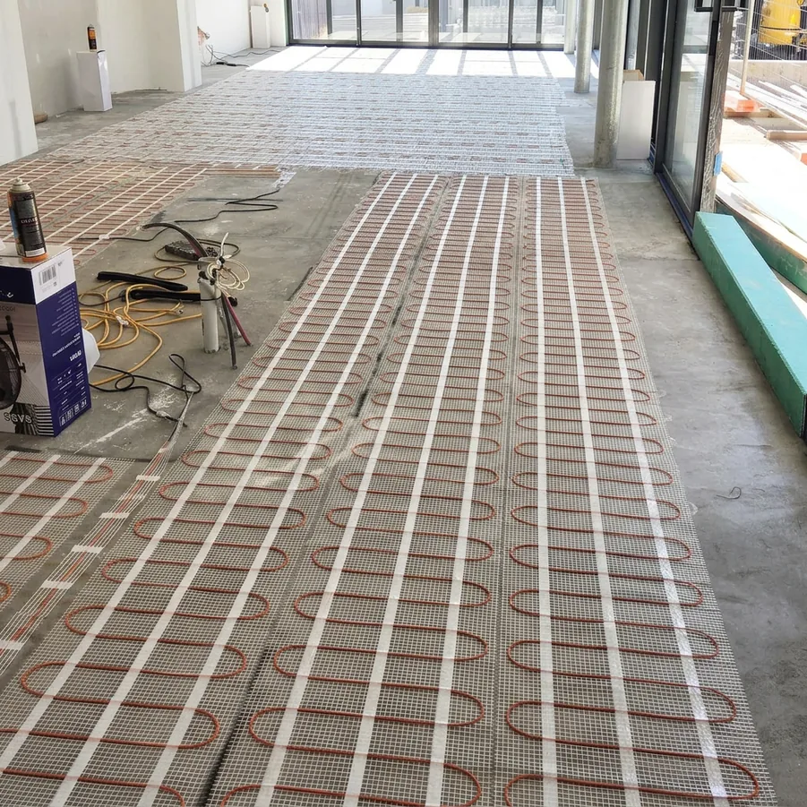 Heating mats unrolled across the floor of a new house, with the cable visible on its mesh backing.